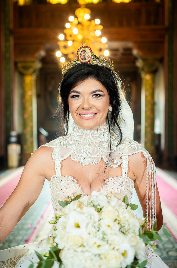 Portret frontal al miresei în timpul slujbei de nuntă într-o biserică din București, purtând o rochie elegantă cu guler înalt și o coroniță pe cap, ținând buchetul de flori albe.