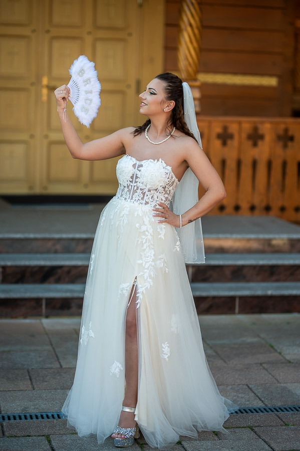 O mireasă zâmbitoare într-o rochie de mireasă albă cu detalii din dantelă, ținând un evantai alb decorat cu cuvântul 'BRIDE', stând pe scările unei clădiri din lemn din București