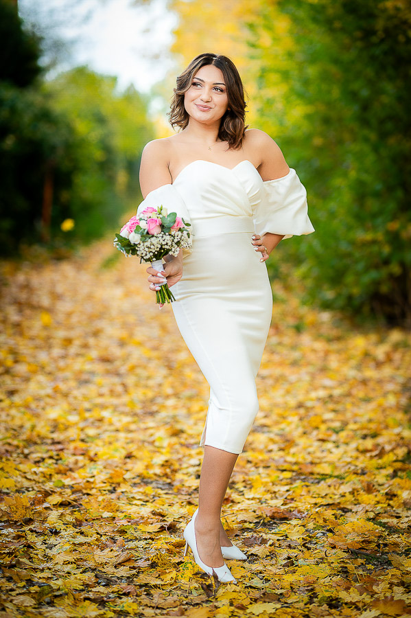 Portret de mireasă realizat de un fotograf de nuntă în București, într-un parc tomnatic cu frunze galbene pe jos. Mireasa poartă o rochie mulată și ține un buchet de trandafiri roz