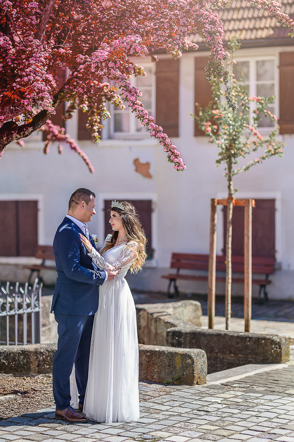 Ședință foto de nuntă în aer liber în București: mirele și mireasa sub un copac cu flori roz vibrante, cu o clădire istorică în fundal, privindu-se tandru.